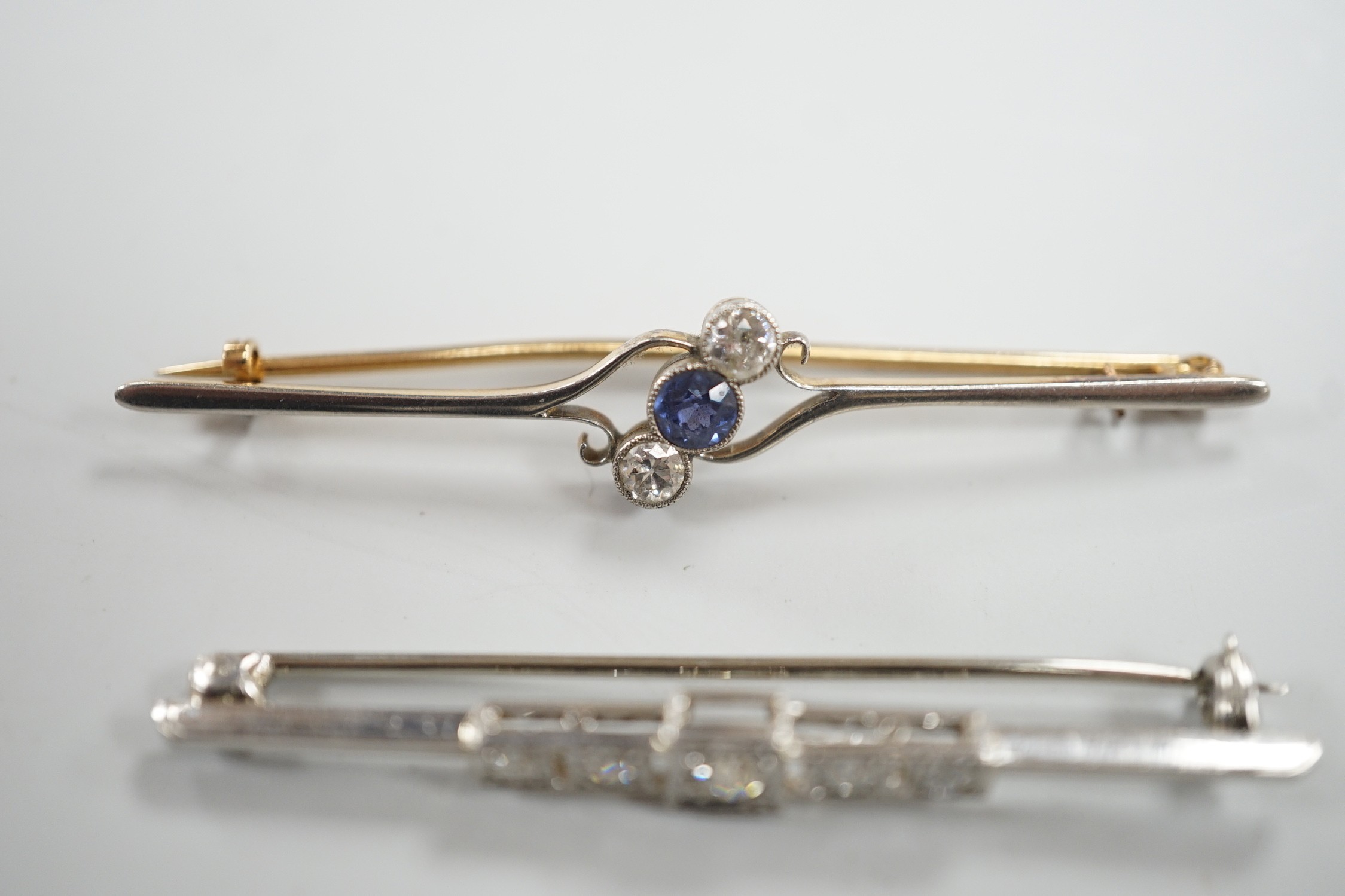 An early to mid 20th century white metal and five stone diamond set bar brooch, 50mm and a similar yellow metal, two stone diamond and single stone sapphire set crossover bar brooch, gross weight 7.1 grams.